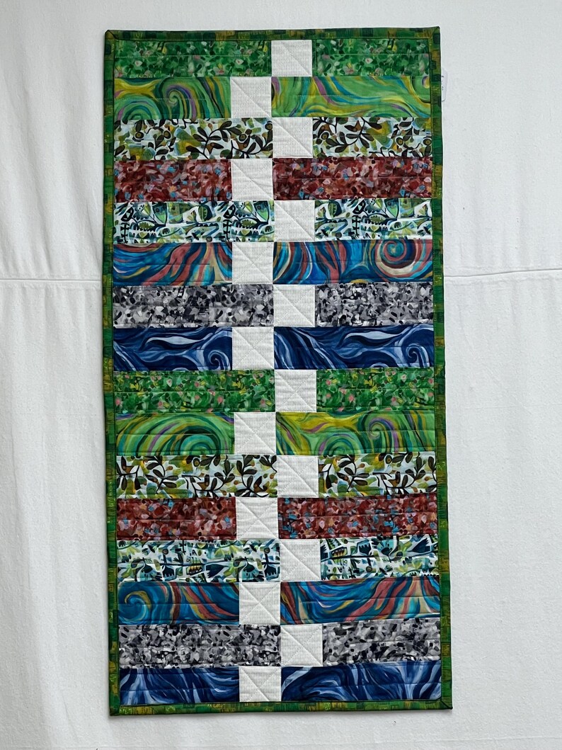 K&ouml;nnte beinhalten: Ein farbenfroher Patchwork-Decken mit einer Vielzahl von Mustern und Farben, darunter Gr&uuml;n, Blau, Rot und Wei&szlig;. Die Decke besteht aus horizontalen Stoffstreifen mit einem wei&szlig;en Quadrat in der Mitte jedes Streifens. Die Decke ist mit einem gr&uuml;nen Rand eingefasst.