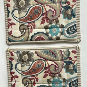 May include: Two beige and brown paisley patterned pot holders with a cream colored border. The pot holders are quilted and have a floral design.