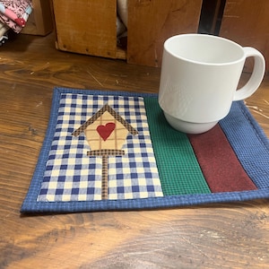 May include: A white mug rests on a patchwork placemat. The placemat features a birdhouse design with a red heart, set against a blue and white checkered pattern, and green and burgundy stripes. The border is blue.