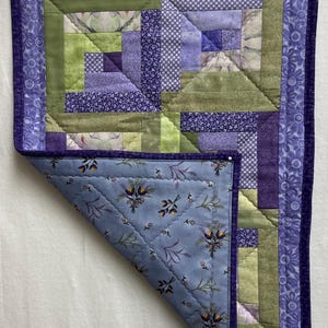 May include: A purple and green patchwork quilt with a floral pattern on the back. The quilt is folded in half and the back is visible.