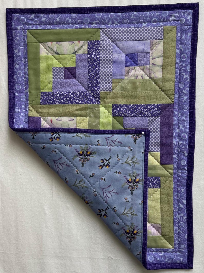 May include: A purple and green patchwork quilt with a floral pattern on the back. The quilt is folded in half and the back is visible.