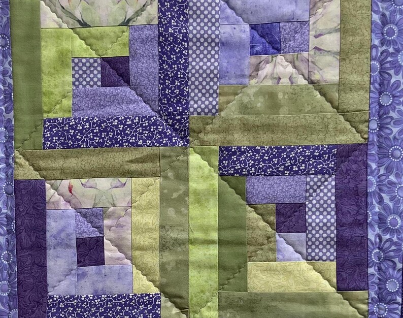 May include: A quilt with a log cabin pattern in shades of green and purple. The quilt is made of different fabrics with floral and geometric designs.