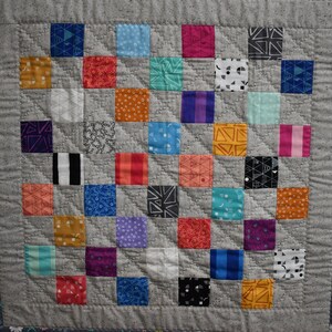 May include: A gray and white quilt with a patchwork design. The quilt is made up of squares of different colors and patterns, including red, blue, green, yellow, and orange. The quilt is bordered with a dark gray fabric with white flowers.