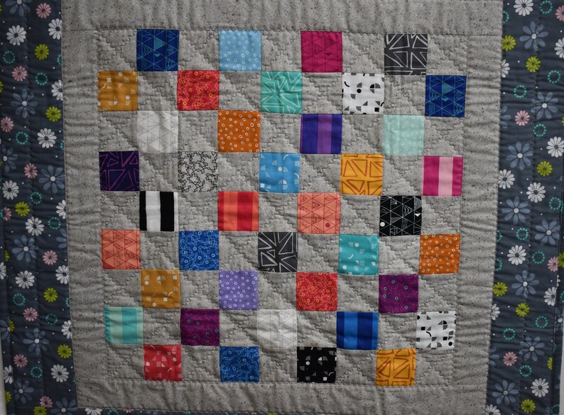 May include: A gray and white quilt with a patchwork design. The quilt is made up of squares of different colors and patterns, including red, blue, green, yellow, and orange. The quilt is bordered with a dark gray fabric with white flowers.