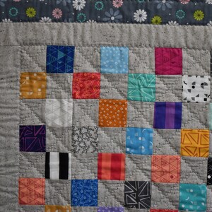 May include: A gray and white quilt with a patchwork design. The quilt is made up of squares of different colors and patterns, including red, blue, green, yellow, and orange. The quilt is edged with a gray and white border.