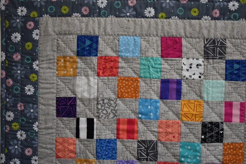 May include: A gray and white quilt with a patchwork design. The quilt is made up of squares of different colors and patterns, including red, blue, green, yellow, and orange. The quilt is edged with a gray and white border.
