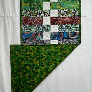 K&ouml;nnte beinhalten: Eine gr&uuml;ne und wei&szlig;e Patchwork-Decke mit geometrischem Muster. Die Decke besteht aus verschiedenen Stoffen mit verschiedenen Mustern, darunter Wirbel, Streifen und florale Motive. Die Decke ist in der H&auml;lfte gefaltet und zeigt eine gr&uuml;ne und gelbe gemusterte R&uuml;ckseite.