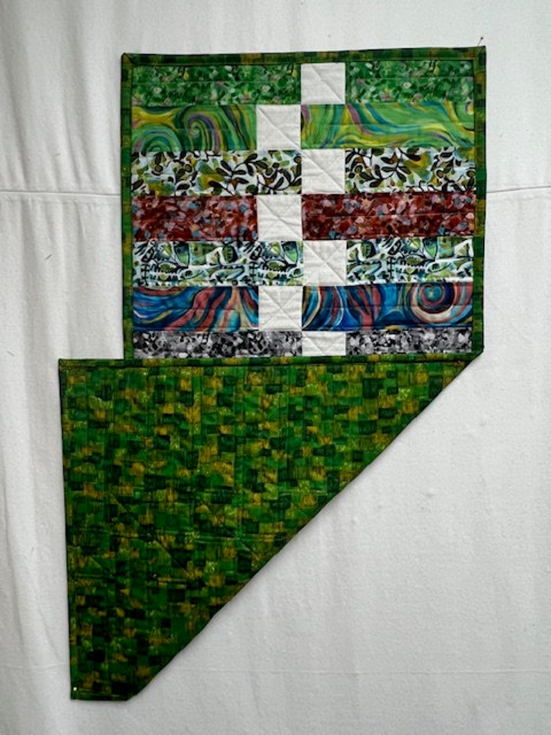 K&ouml;nnte beinhalten: Eine gr&uuml;ne und wei&szlig;e Patchwork-Decke mit geometrischem Muster. Die Decke besteht aus verschiedenen Stoffen mit verschiedenen Mustern, darunter Wirbel, Streifen und florale Motive. Die Decke ist in der H&auml;lfte gefaltet und zeigt eine gr&uuml;ne und gelbe gemusterte R&uuml;ckseite.