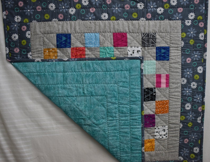 May include: A teal blue quilted fabric with a gray and white patterned fabric border. The gray fabric has a patchwork design with squares of various colors and patterns.