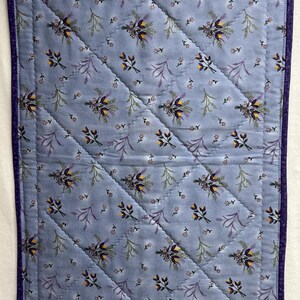 May include: A blue quilted fabric with a floral pattern of purple and yellow flowers. The fabric has a diamond pattern of stitching.