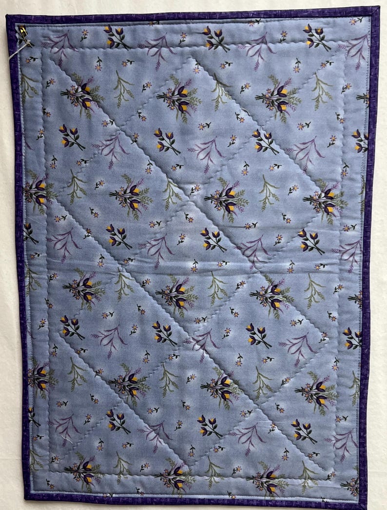 May include: A blue quilted fabric with a floral pattern of purple and yellow flowers. The fabric has a diamond pattern of stitching.