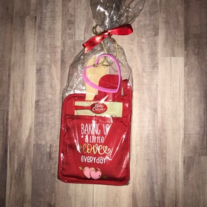 Op de afbeelding: Een rode ovenwant met de tekst "Baking up a little love everyday" in gouden letters en twee roze harten. De want is verpakt met een houten lepel en een roze hartvormige koekjesuitsteker.