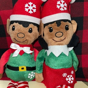 Può includere: Due bambole di elfo di peluche con gambe a strisce rosse e bianche, abiti da elfo verdi e bianchi e cappelli da Babbo Natale rossi con fiocchi di neve bianchi. Le bambole hanno la pelle marrone e i capelli neri.