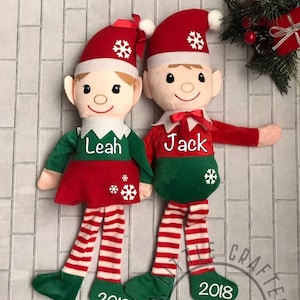 Può includere: Due elfi di Natale personalizzati con abiti rossi e verdi, fiocchi di neve bianchi e i nomi "Leah" e "Jack" ricamati sulle loro camicie. Gli elfi indossano calze a righe e hanno l'anno "2018" ricamato sui loro piedi.