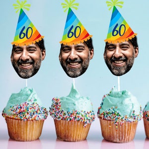 May include: Three cupcakes with blue frosting and colorful sprinkles. Each cupcake has a birthday hat with a face on it. The hats are rainbow colored and say "60" on them.