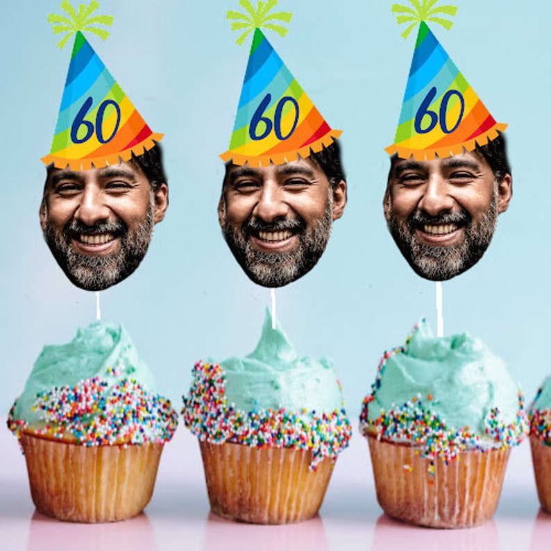 May include: Three cupcakes with blue frosting and colorful sprinkles. Each cupcake has a birthday hat with a face on it. The hats are rainbow colored and say "60" on them.