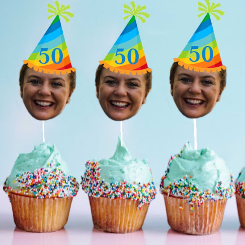 May include: Three cupcakes with blue frosting and colorful sprinkles topped with a face cutout on a stick wearing a rainbow party hat with the number 50 on it.
