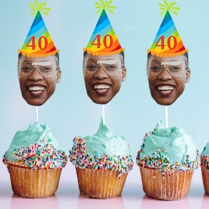 May include: Three cupcakes with blue frosting and colorful sprinkles topped with a face cutout on a stick. The face cutouts are wearing rainbow party hats with the number "40" on them.