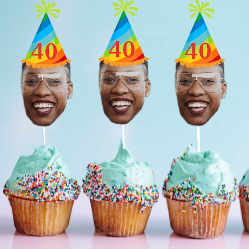 May include: Three cupcakes with blue frosting and colorful sprinkles topped with a face cutout on a stick. The face cutouts are wearing rainbow party hats with the number "40" on them.
