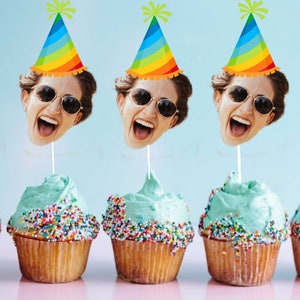 May include: Three cupcakes with blue frosting and colorful sprinkles topped with a face cutout wearing a rainbow party hat.