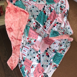 May include: A pink and white fleece blanket with a patchwork design featuring a variety of floral patterns, hearts, and the text "Nursing heart" repeated throughout. The blanket has a soft, fuzzy texture and is perfect for cuddling up on the couch.