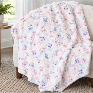 May include: A plush white throw blanket featuring a repeating pattern of embossed paw prints in soft shades of pink and light blue. The fabric has a textured, raised surface with a soft, fuzzy appearance draped over a chair.