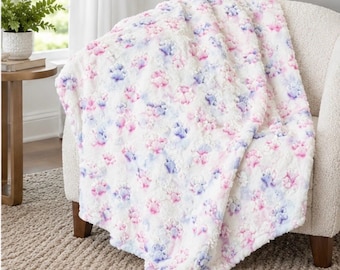 May include: A plush white throw blanket featuring a repeating pattern of embossed paw prints in soft shades of pink and light blue. The fabric has a textured, raised surface with a soft, fuzzy appearance draped over a chair.