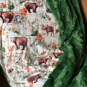 May include: A green plush throw blanket with a white and brown patterned side featuring a variety of woodland animals, including bears, deer, raccoons, and moose. The pattern includes leaves, trees, and the words "Moose Lodge".