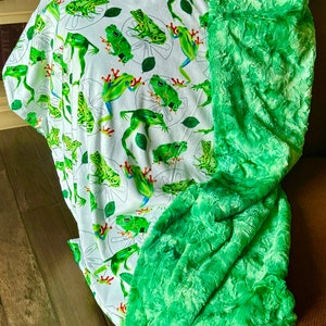 May include: A soft throw blanket featuring a white background with a repeating pattern of green frogs and lily pads. One side of the blanket is made of a smooth fabric with the frog print, while the other side is a textured, plush bright green fabric.