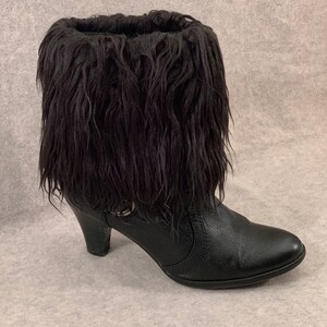 May include: Black leather ankle boot with a high heel and a fluffy black fur collar. The boot features a small buckle detail on the side. The heel is approximately 3 inches high.