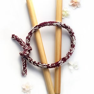 May include: A burgundy and white braided bracelet, knotted at one end, with two wooden dowels behind it. The image is set against a white background, with small, delicate flowers. The bracelet is a handcrafted item.