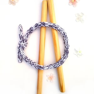 May include: Two wooden dowels with a grey and white braided rope wrapped around them. The rope is tied in a knot at the top. There are several small white flowers scattered around the dowels.