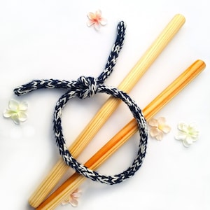May include: Two wooden dowels with a black and white knitted rope tied around them. The rope is tied in a knot and the dowels are crossed over each other. There are pink flowers scattered around the dowels.