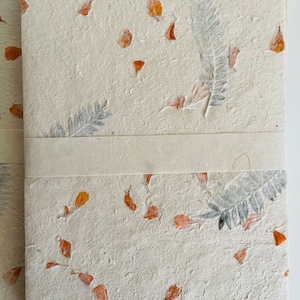 May include: A handmade notebook with a cream-colored cover featuring pressed orange and gray botanicals. The notebook has a white ribbon bookmark.