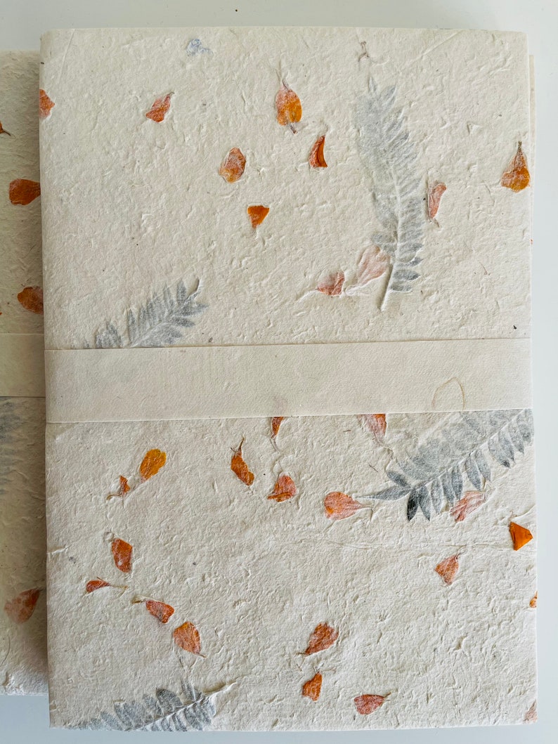 May include: A handmade notebook with a cream-colored cover featuring pressed orange and gray botanicals. The notebook has a white ribbon bookmark.