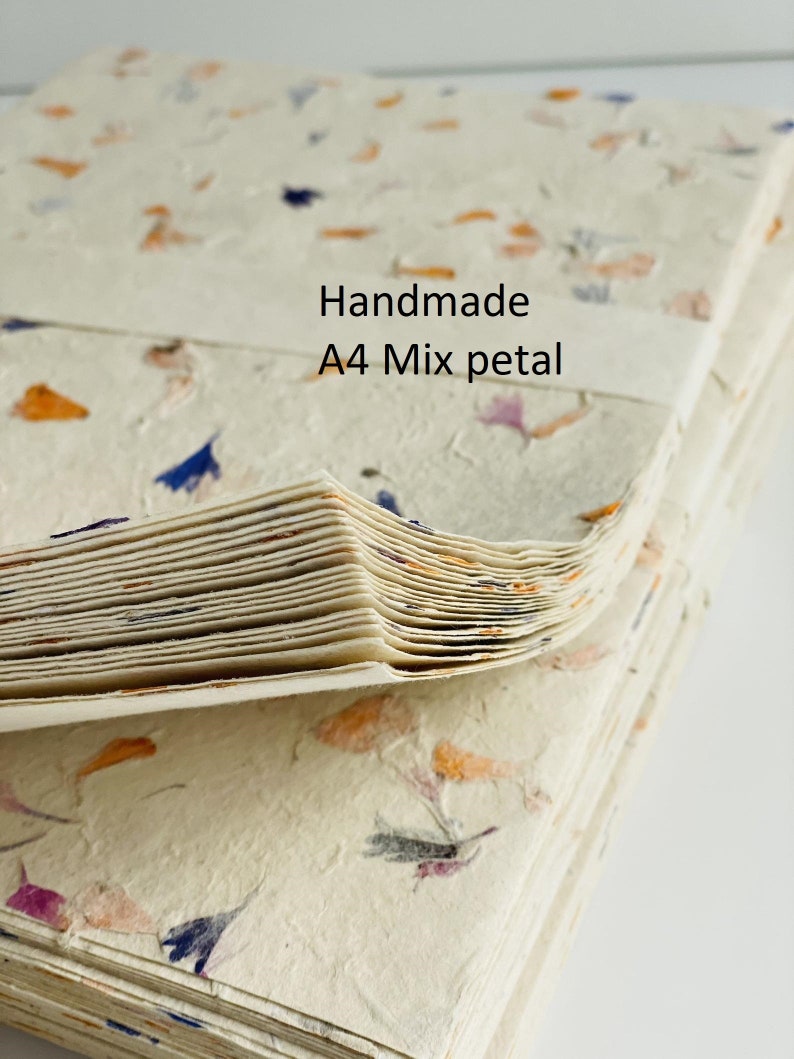 May include: A stack of handmade A4 paper sheets with mixed flower petals embedded in the paper. The paper is cream colored with orange, purple, and blue flower petals.