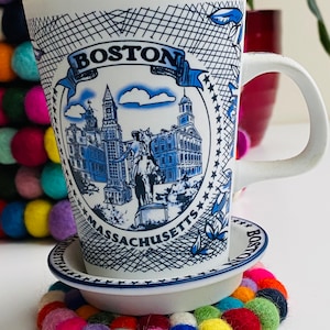 May include: White ceramic mug with a blue and white design featuring the text "BOSTON MASSACHUSETTS" and a depiction of the Boston skyline. The mug is sitting on a white saucer and a colorful felt coaster.