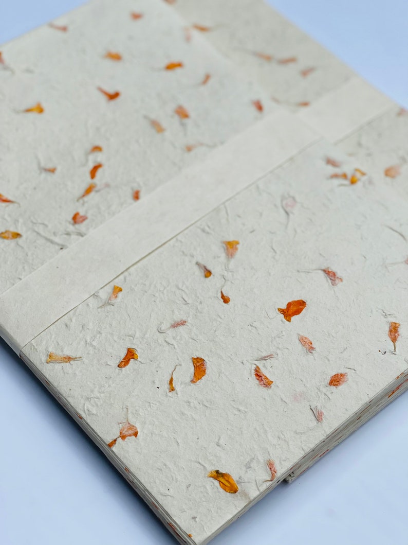 May include: A handmade journal with a white cover and a textured surface. The cover is decorated with small, orange flower petals.