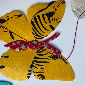K&ouml;nnte beinhalten: Ein gelber Schmetterling mit schwarzen Streifen und einer roten Schleife mit goldenen Sternen und wei&szlig;en Punkten. Der Schmetterling ist aus Papier und h&auml;ngt an einer Schnur.