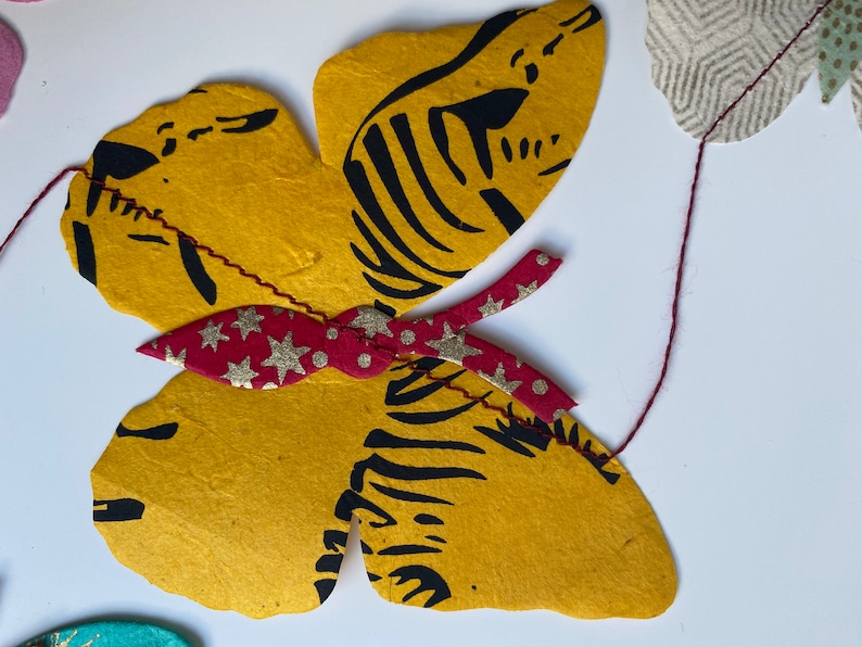 K&ouml;nnte beinhalten: Ein gelber Schmetterling mit schwarzen Streifen und einer roten Schleife mit goldenen Sternen und wei&szlig;en Punkten. Der Schmetterling ist aus Papier und h&auml;ngt an einer Schnur.