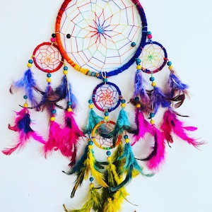 May include: A colorful dreamcatcher with a rainbow-colored outer ring and smaller inner rings. It features a variety of feathers in shades of purple, pink, teal, and yellow.