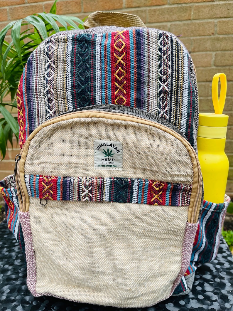 May include: A beige hemp backpack with colourful striped fabric accents. The backpack has a zipped pocket on the front and a label that reads "Himalayan Hemp THC Free Made in Nepal".