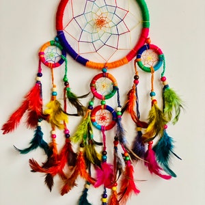 May include: A colorful dream catcher with multiple hoops and feathers. The main hoop is decorated with rainbow colored yarn and has a white web inside. The smaller hoops are also decorated with colorful yarn and beads. The feathers are a variety of colors, including red, orange, yellow, green, blue, and purple.