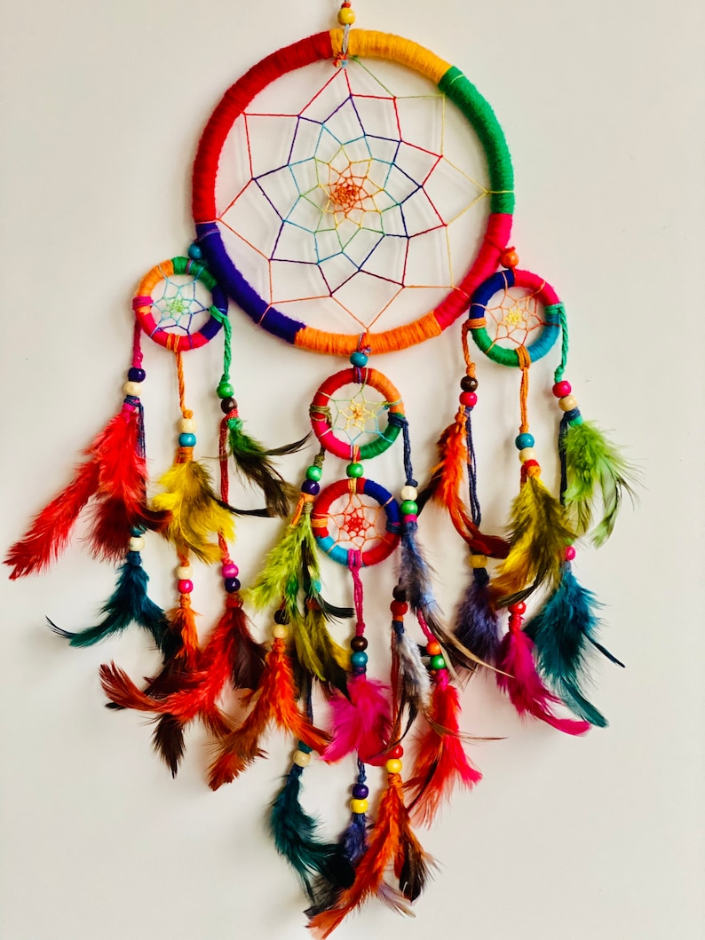 May include: A colorful dream catcher with multiple hoops and feathers. The main hoop is decorated with rainbow colored yarn and has a white web inside. The smaller hoops are also decorated with colorful yarn and beads. The feathers are a variety of colors, including red, orange, yellow, green, blue, and purple.