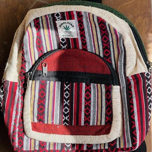 May include: A red, black, and white striped hemp backpack with a brown leather strap and a label that reads "Himalayan Hemp The Free Made in Nepal".