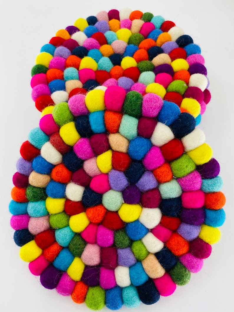 May include: Two round felt coasters made of colorful wool balls. The coasters are arranged in a stack with the top coaster slightly offset from the bottom coaster. The coasters are a mix of bright colors, including red, orange, yellow, green, blue, purple, pink, and white.