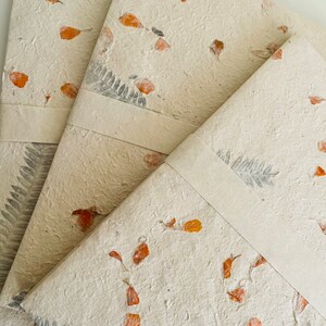May include: Three handmade journals with a natural, off-white paper cover. The covers are decorated with pressed flowers and leaves in shades of orange and brown.