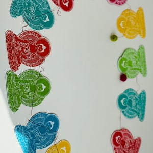 K&ouml;nnte beinhalten: Eine Girlande aus Papierschnitten von Buddha-Figuren in verschiedenen Farben, darunter Rot, Blau, Gr&uuml;n und Gelb. Die Figuren sind durch eine d&uuml;nne Schnur verbunden und in einem Kreis angeordnet.