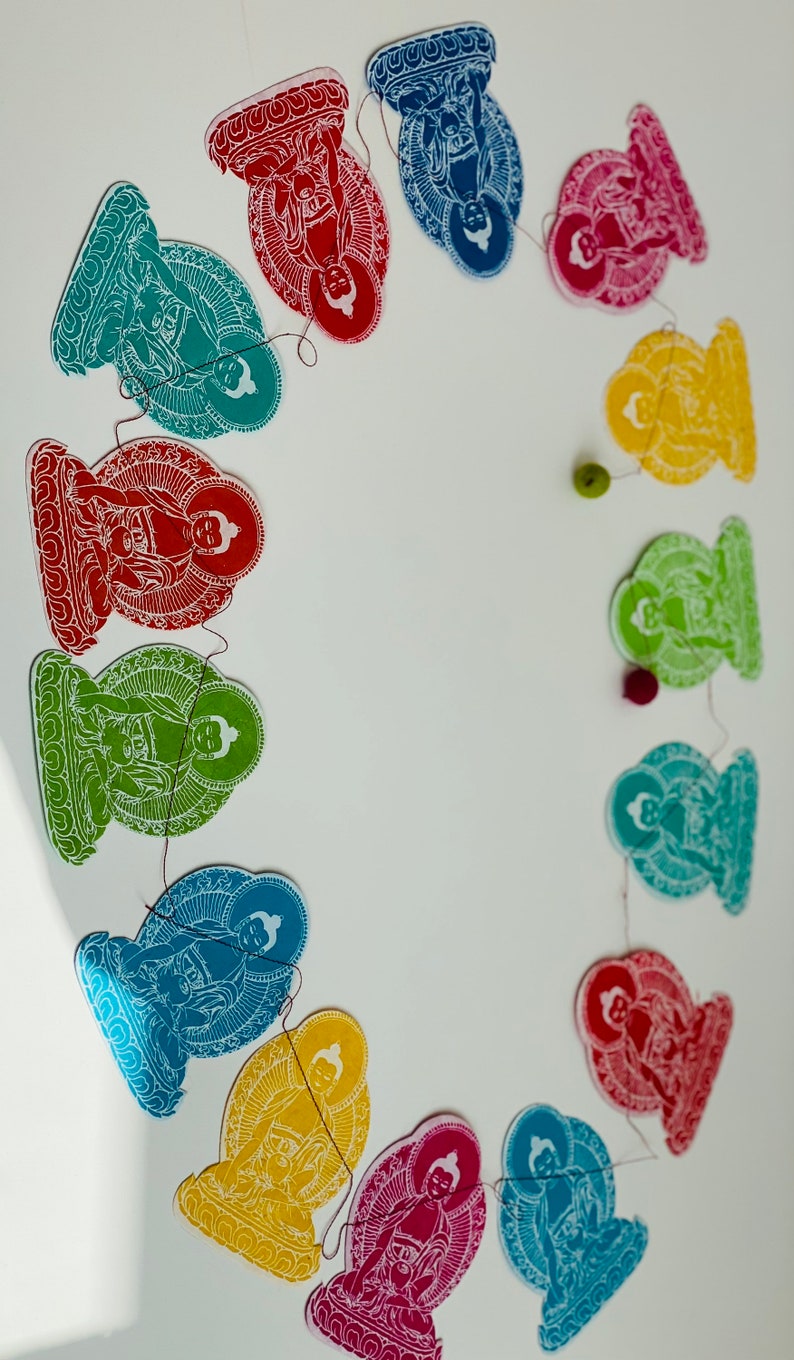 K&ouml;nnte beinhalten: Eine Girlande aus Papierschnitten von Buddha-Figuren in verschiedenen Farben, darunter Rot, Blau, Gr&uuml;n und Gelb. Die Figuren sind durch eine d&uuml;nne Schnur verbunden und in einem Kreis angeordnet.