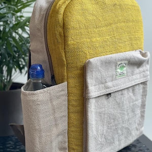 May include: A yellow and beige backpack made from textured fabric. The backpack features a large main compartment, a side pocket holding a water bottle, and a front pocket. The side and front pockets are beige, while the main body is yellow. It has a zip closure.